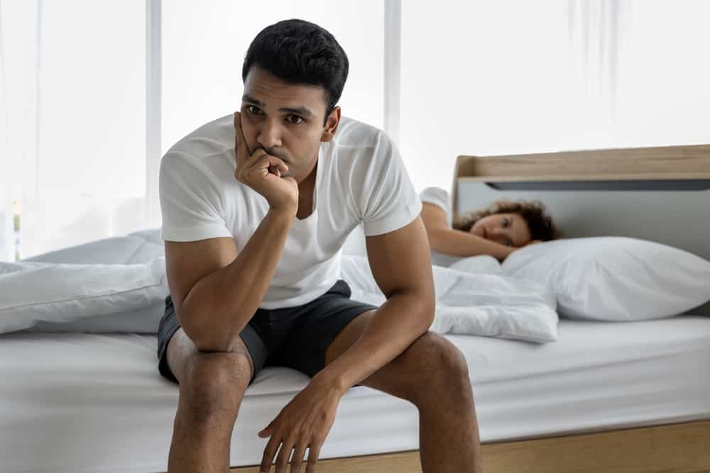 Upset depressed young man sitting on the edge of bed against his wife lying on the bed. Relationship difficulties.